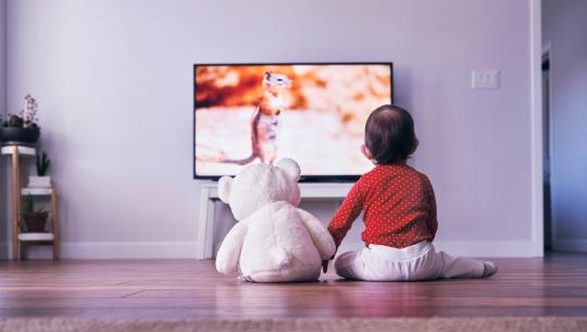Image of a kid sitting with a teddy bear watching television.