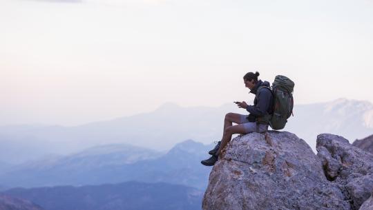 Image of a hiker sitting on a rocky cliff using a mobile phone.