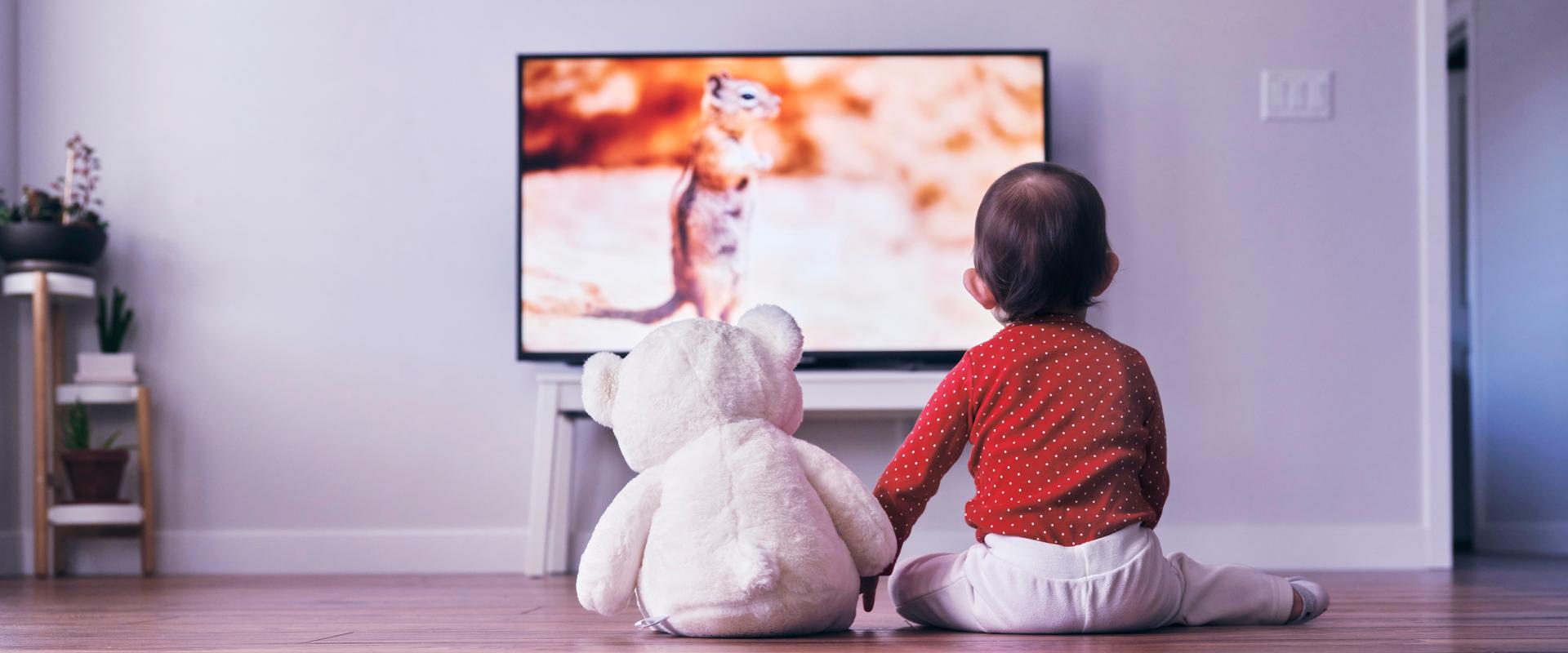 Image of a kid sitting with a teddy bear watching television.