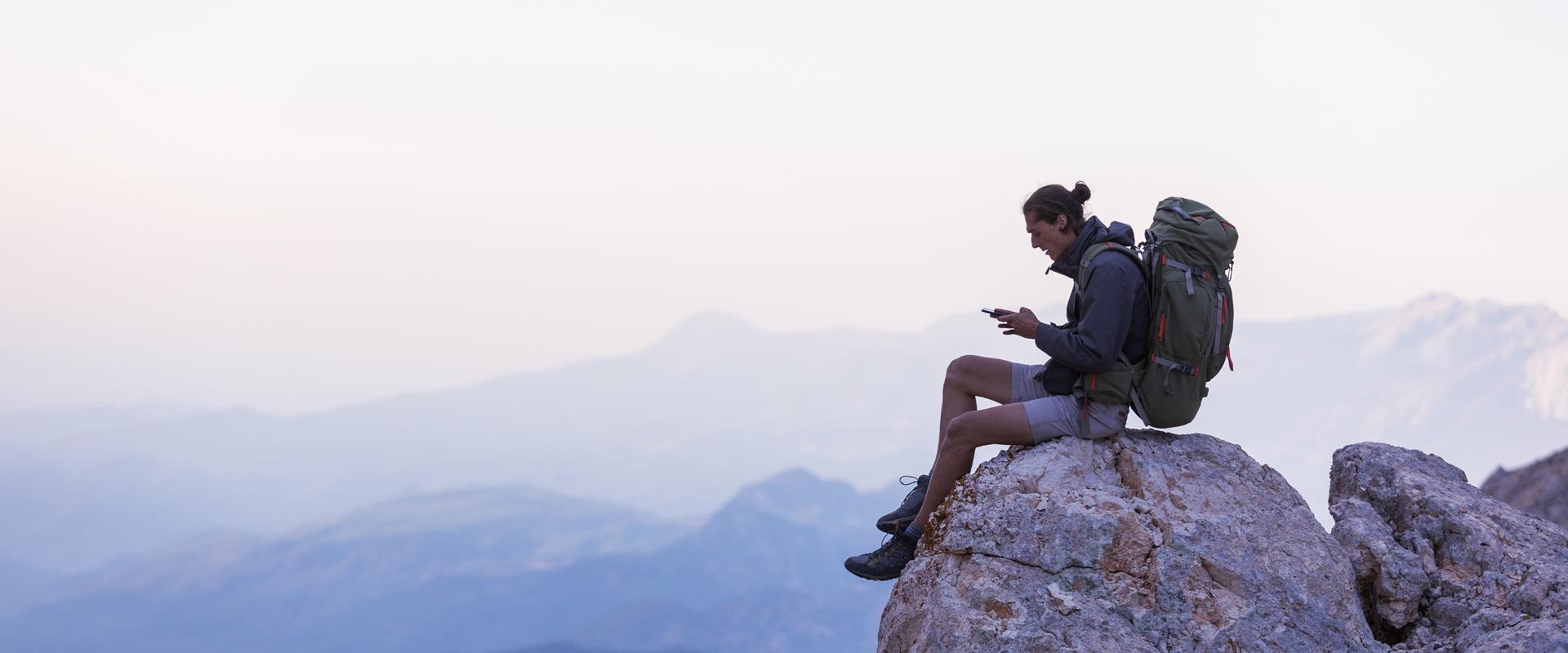 Image of a hiker sitting on a rocky cliff using a mobile phone.