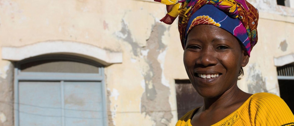 Image of a woman wearing a colorful headwrap in front of an old, weathered building.