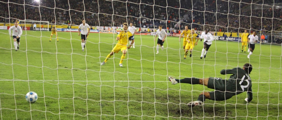 Image of goalkeeper diving as the ball heads toward the net during a football match.