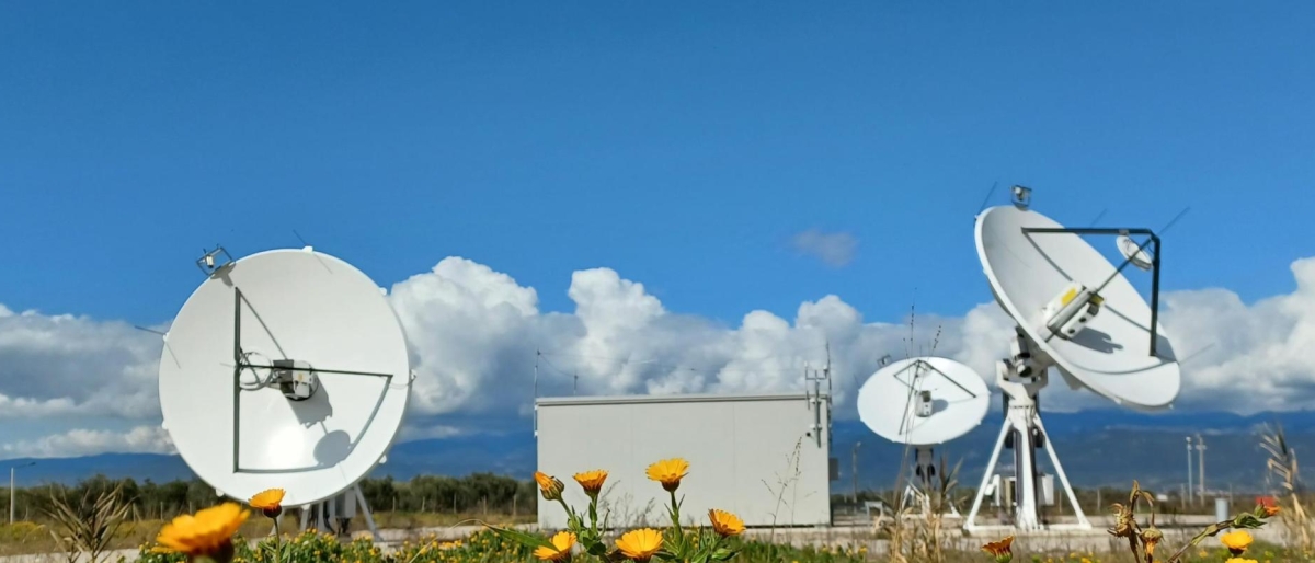 Image of satellite dishes in a field with yellow flowers under a bright blue sky.