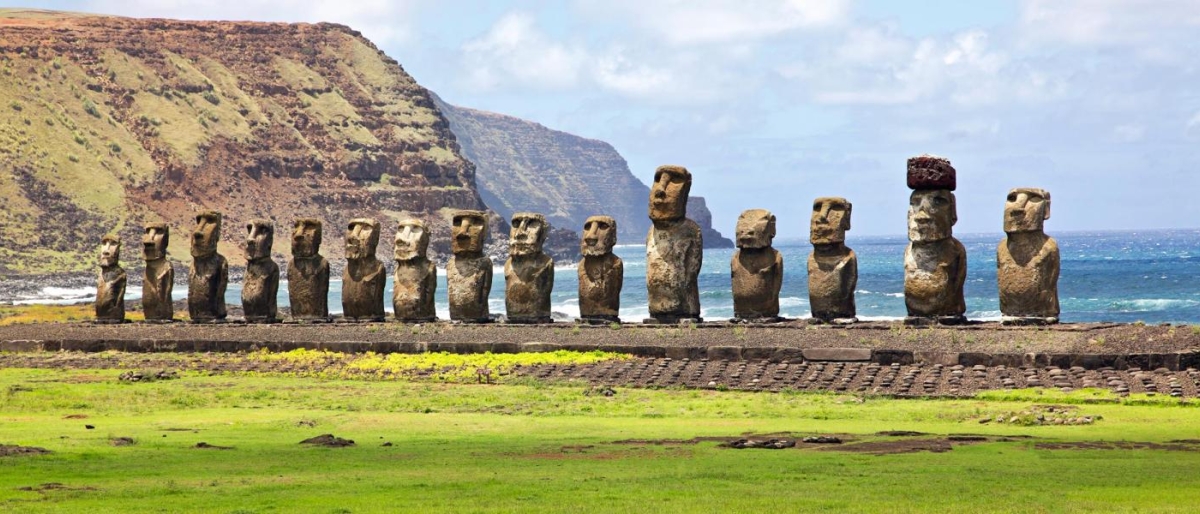 Image shows Row of Moai statues with ocean cliffs in the background.