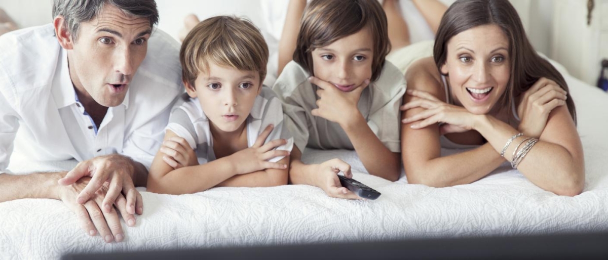 Image of a family lying on a bed together while watching TV.