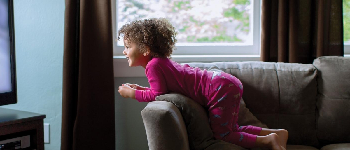 Image of a child in pajamas kneeling on a couch watching TV.