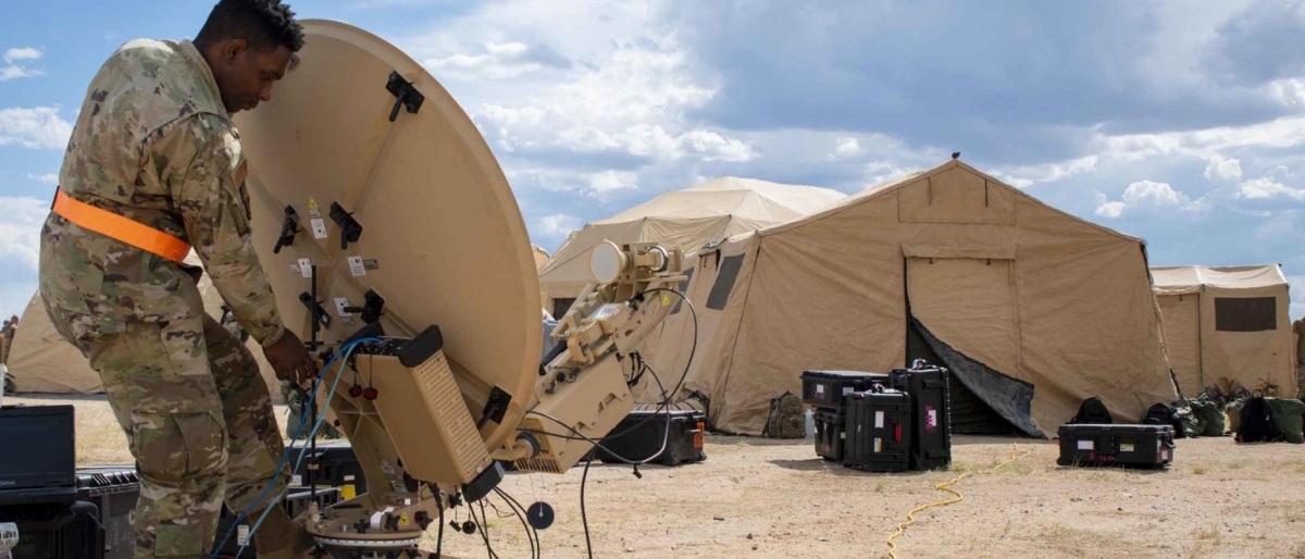 Image of a soldier setting up a portable satellite dish beside field tents.