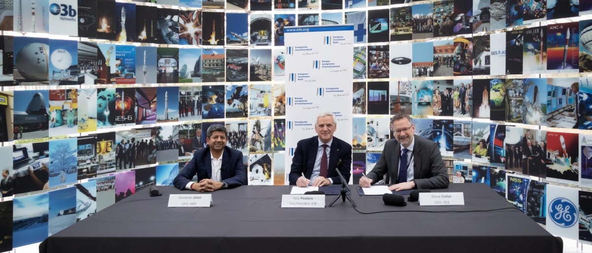 Image of three representatives signing documents at a table in front of a wall of technology and space‑related images.