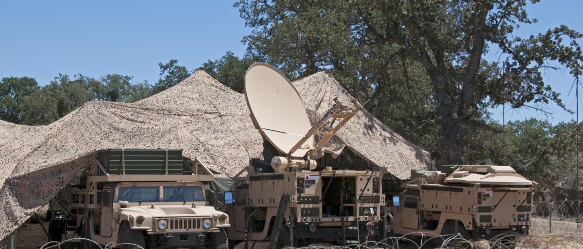 Image of a military vehicles with a large satellite dish set up under camouflage netting.