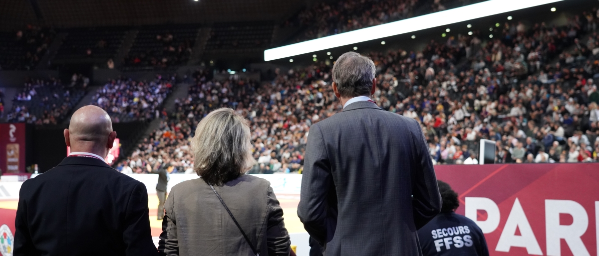People standing at the edge of a judo arena facing a large, crowded audience.