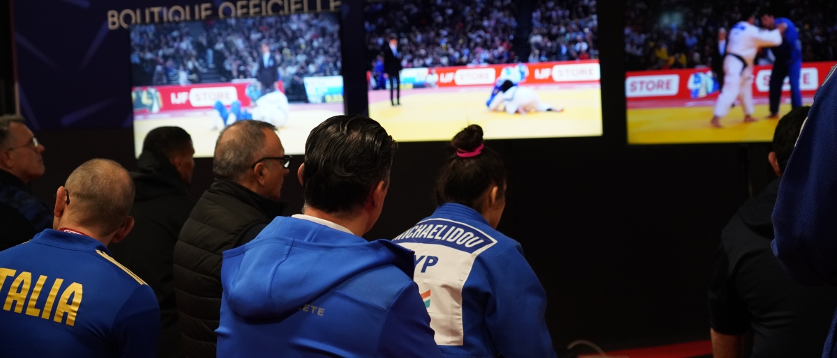Athletes watching a judo match on multiple screens backstage.