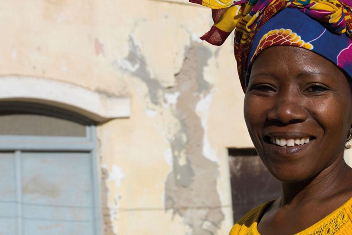 Image of a woman wearing a colorful headwrap in front of an old, weathered building.