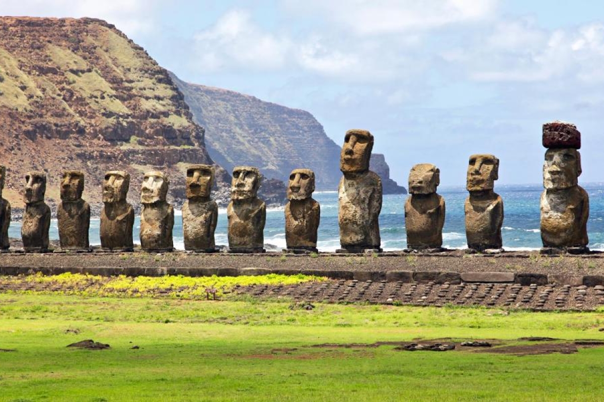 Image shows Row of Moai statues with ocean cliffs in the background.