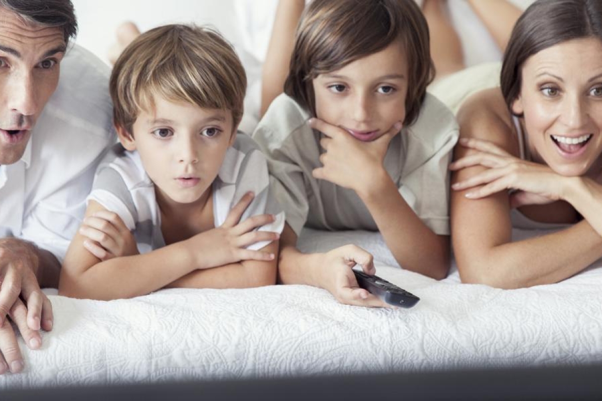 Image of a family lying on a bed together while watching TV.