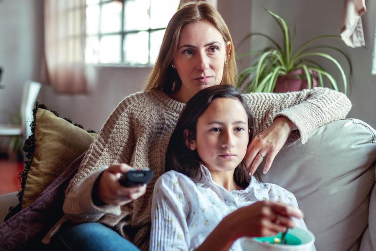 Image of an adult and a kid sitting on the couch watching television.