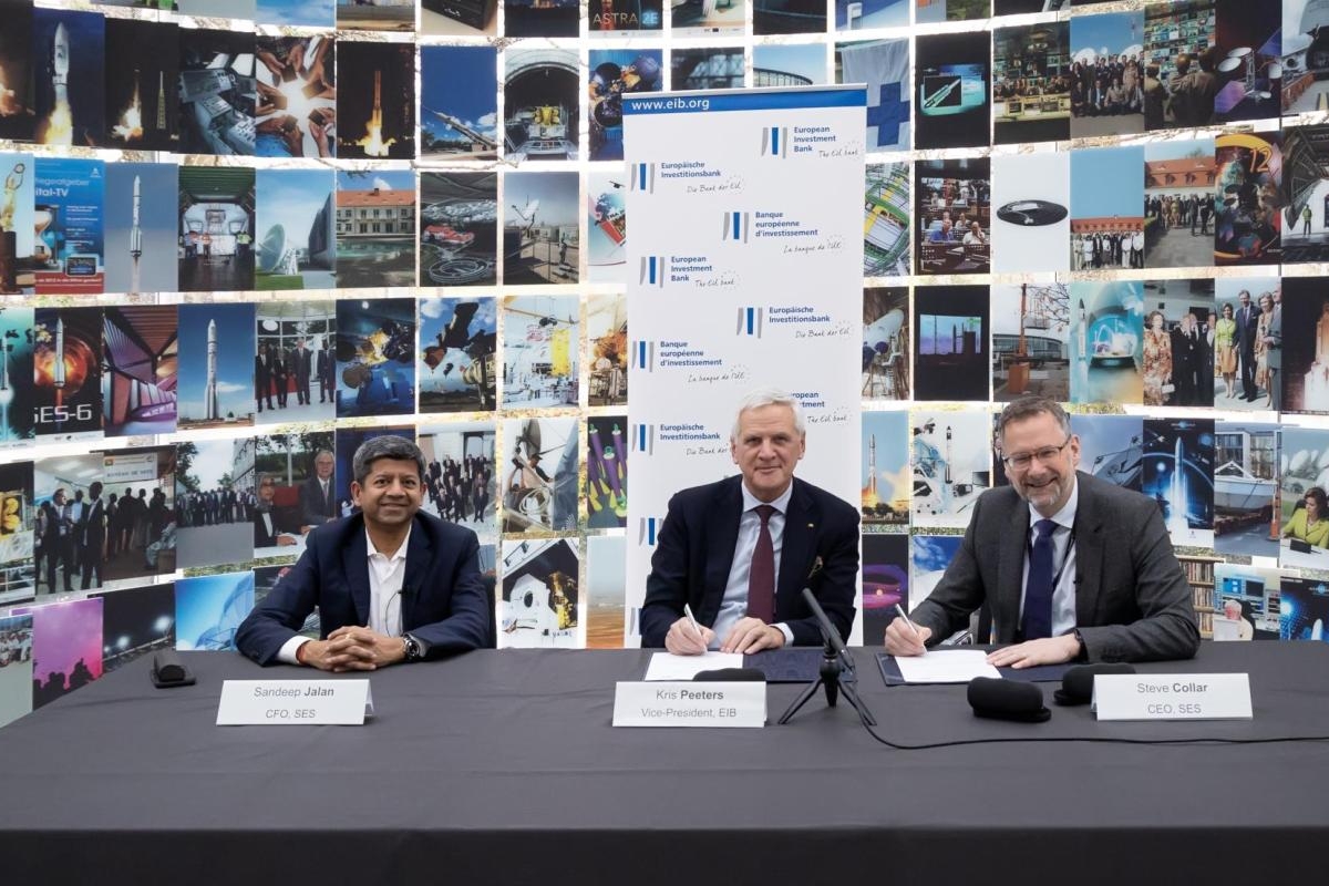 Image of three representatives signing documents at a table in front of a wall of technology and space‑related images.