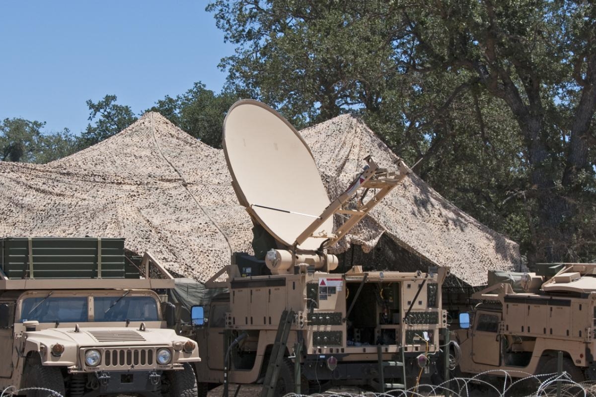 Image of a military vehicles with a large satellite dish set up under camouflage netting.