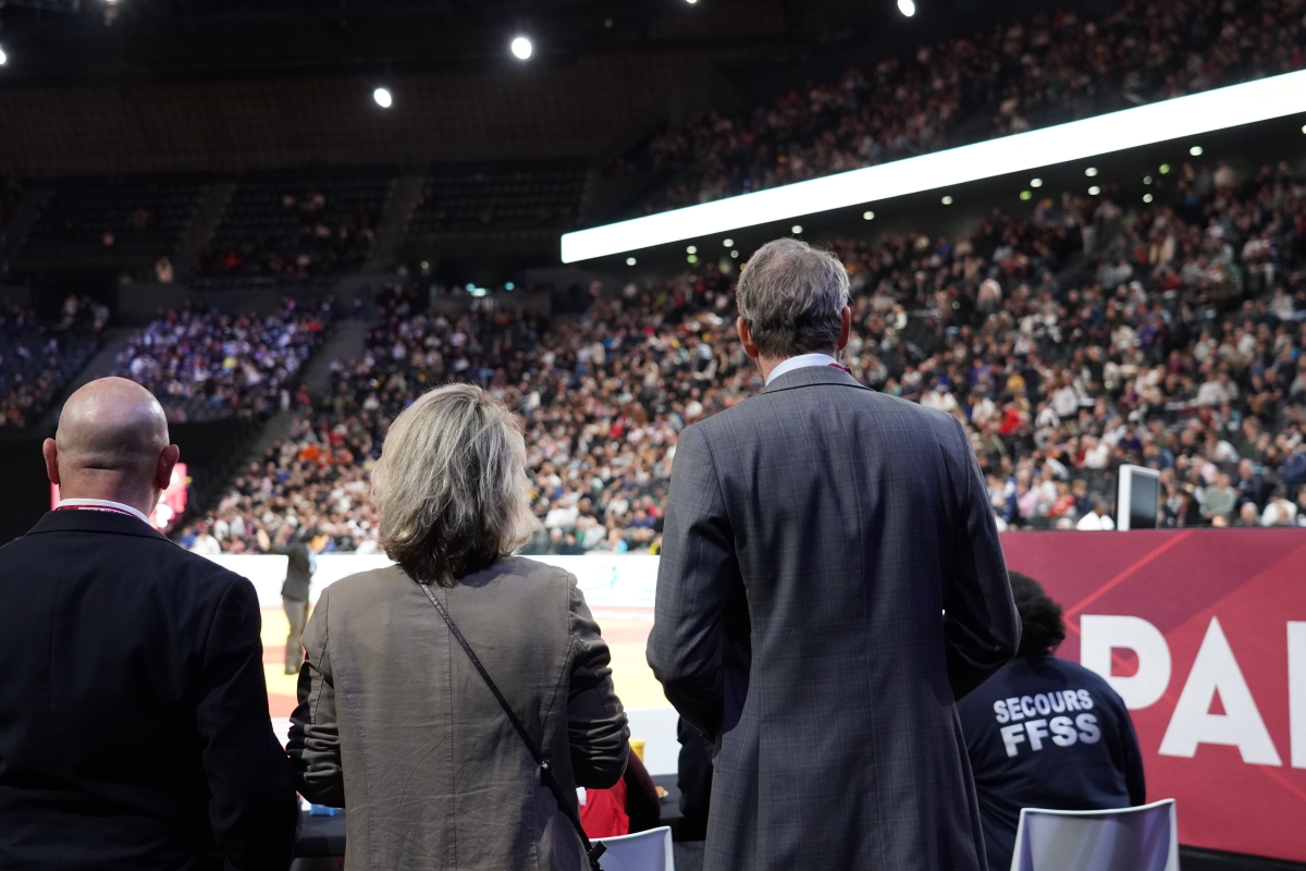 People standing at the edge of a judo arena facing a large, crowded audience.
