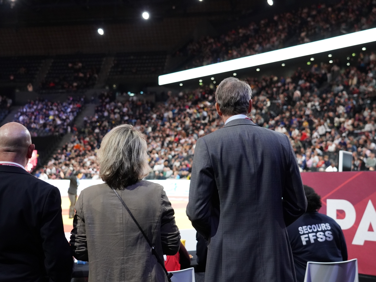People standing at the edge of a judo arena facing a large, crowded audience.