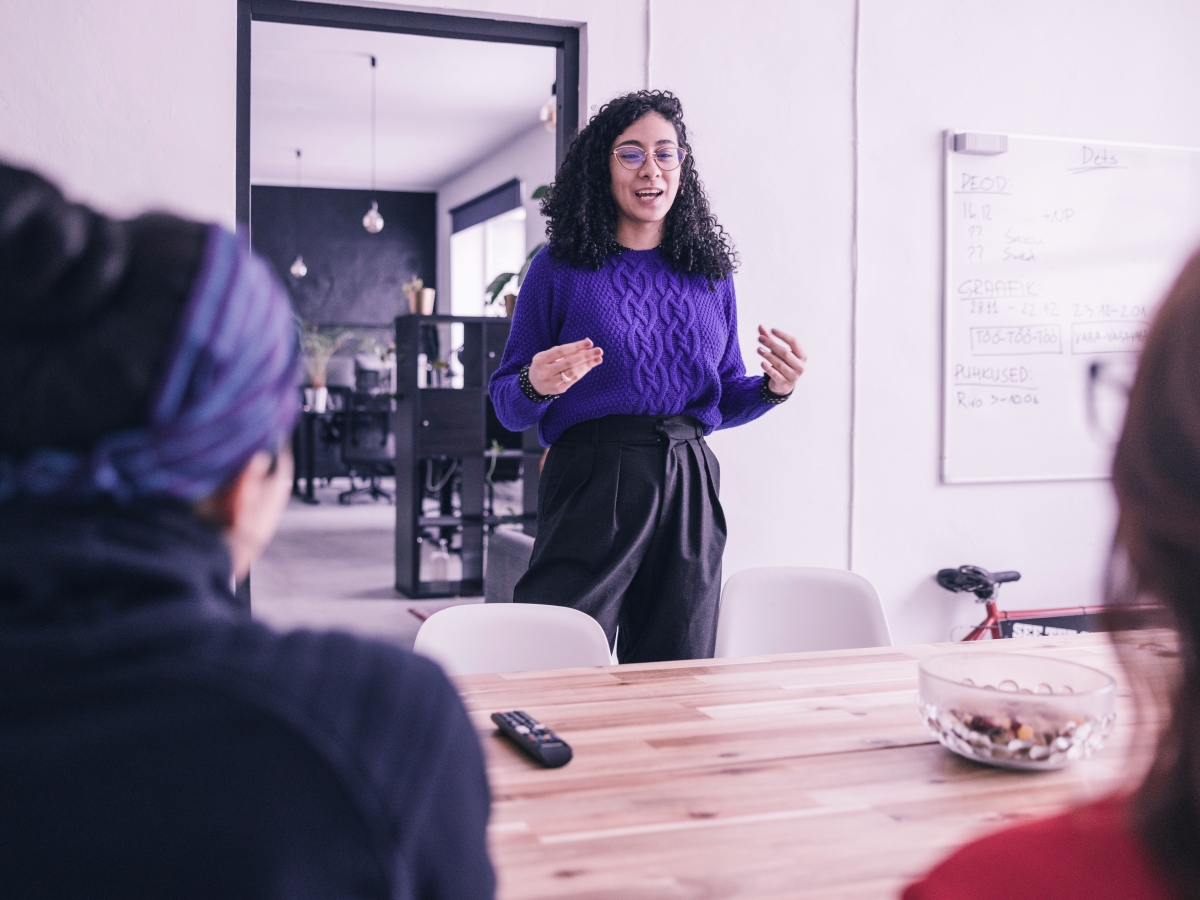 Person presenting to colleagues in a meeting room, standing near a whiteboard.
