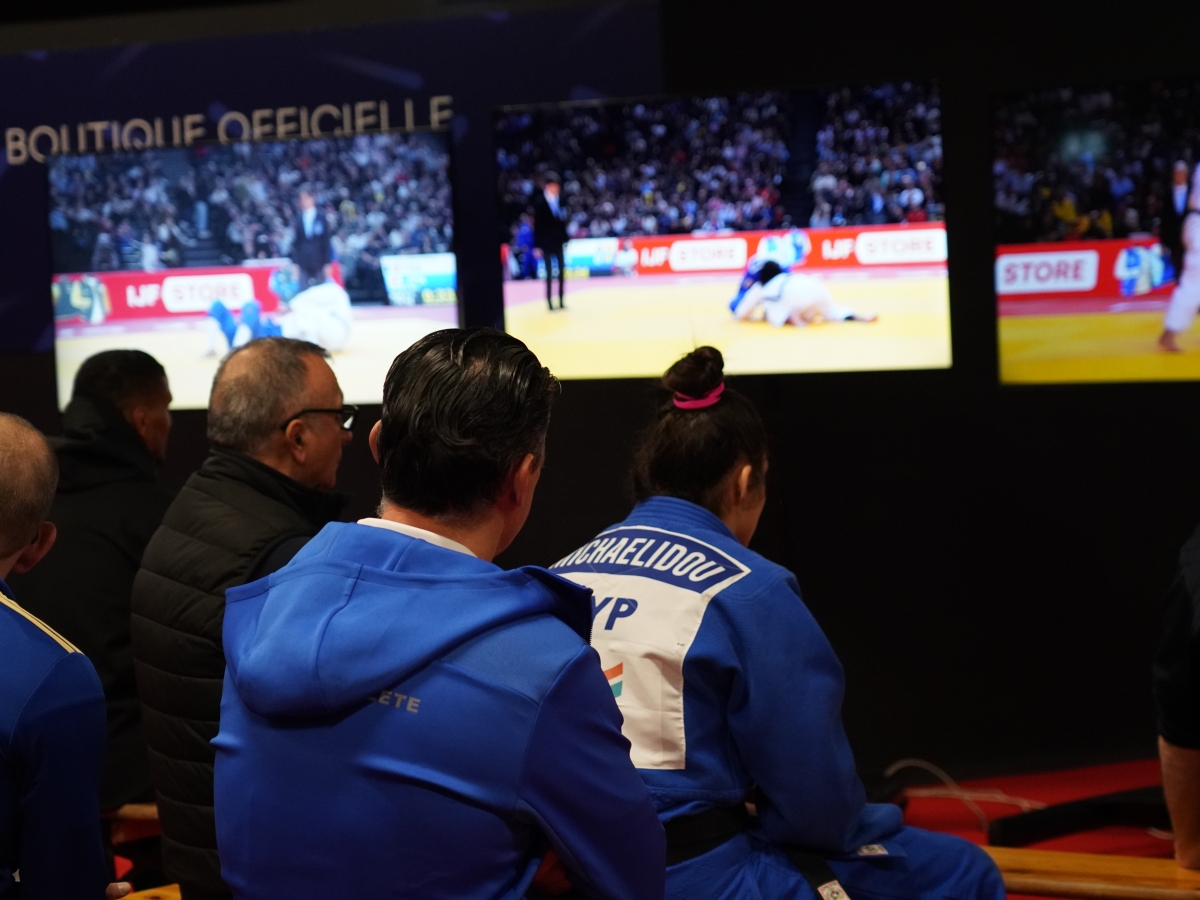 Athletes watching a judo match on multiple screens backstage.