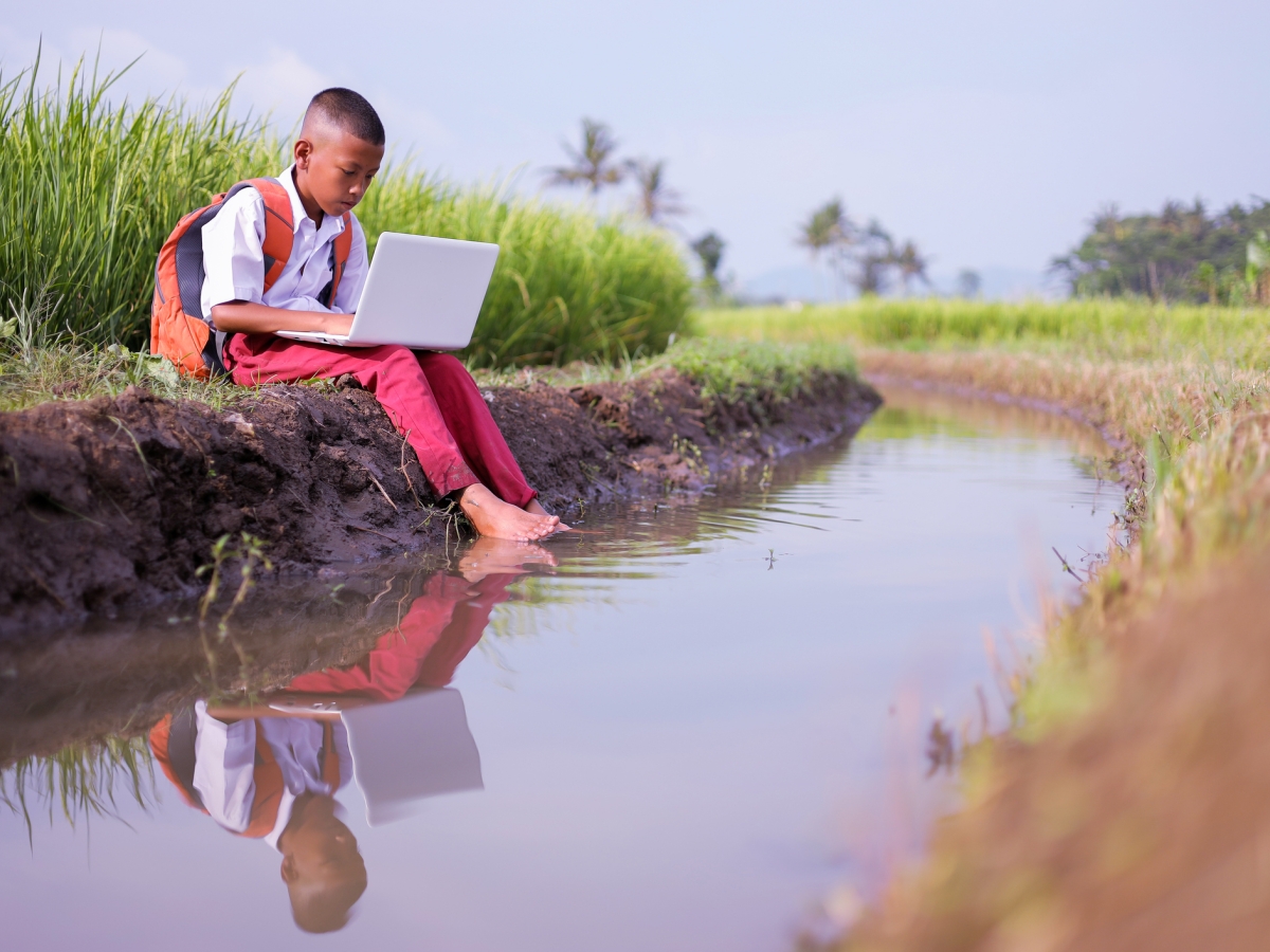 Image of a kid studying using a laptop near a stream in a rural area.